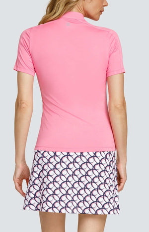Back view of a woman wearing a light pink short sleeve golf top. There are inserts on the top of the sleeves. She is also wearing a golf skirt with a circular geometric pattern with light pink and dark navy blue on a white background.