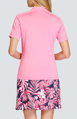 Back view of a woman wearing a light pink short sleeve golf top. There are inserts on the top of the sleeves. She is also wearing a golf skirt with a leaf pattern in the same light pink and a dark navy blue background.