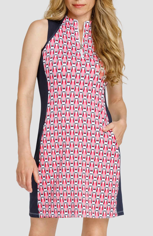 Front view of a woman wearing a sleeveless golf dress with a quarter-zip neck opening. The print is a geometric pattern of 6-sided gemstones in a light pink color on a white background. There are side and shoulder inserts in a dark navy blue color. The woman has her hand in one of the pockets.
