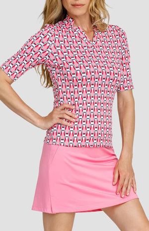 Three-quarter view of a woman wearing a mid-length sleeve golf top with a star collar neckline. The print is a geometric pattern of 6-sided gemstones in a light pink color on a white background. She is also wearing a light pink golf skort.