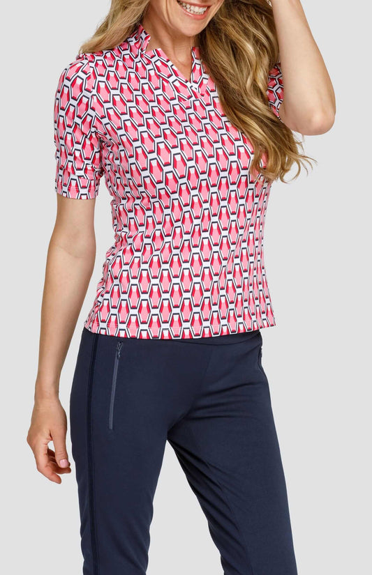 Front view of a woman wearing a mid-length sleeve golf top with a star collar neckline. The print is a geometric pattern of 6-sided gemstones in a light pink color on a white background. She is also wearing dark navy blue pants with zipper pockets.