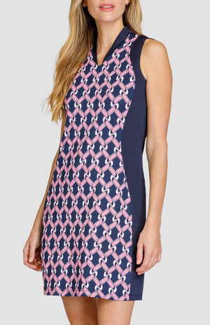 Three-quarter view of a woman wearing a sleeveless golf dress with an open v-neck collar. The print is a light pink and white chain geometric pattern on a dark navy blue background. There are dark navy blue side inserts from the shoulder to the hem. 