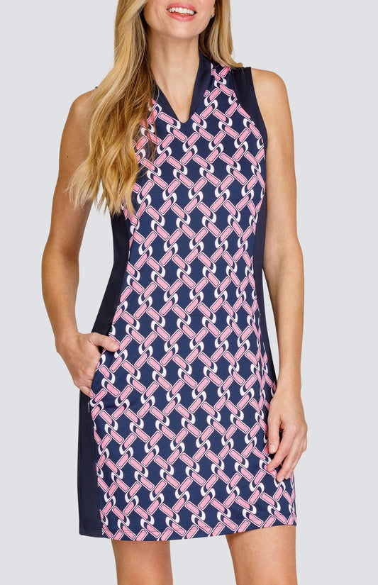 Front view of a woman wearing a sleeveless golf dress with an open v-neck collar. The print is a light pink and white chain geometric pattern on a dark navy blue background. There are dark navy blue side inserts from the shoulder to the hem. She has a hand in one of the pockets.