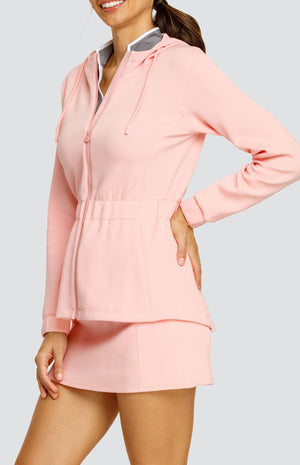 Model wearing a light pink hooded jacket with a cinched waist and drawstring hood. Paired with a matching skort with drawstring waistband, and a gray top with white accents.